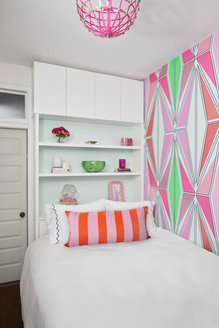 Colorful globe light hangs above bedroom lined with graphic pink, green and orange wallpaper. Open shelves behind the bed are lined with decorative objects. One purple and orange striped pillow placed on neatly made bed.