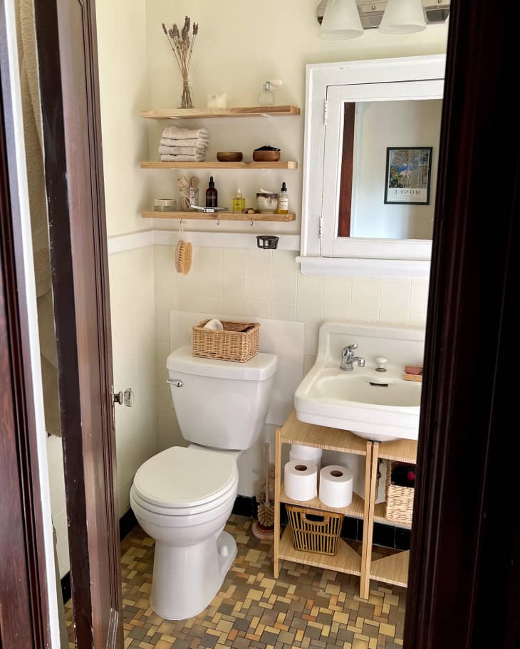 bathroom with brown small tile floor, pale cream walls with white trim, white sink with wood shelves underneath holding toilet paper, bins, 3 floating wood shelves over the toilet with washcloths, toiletries, small vase with dried flowers