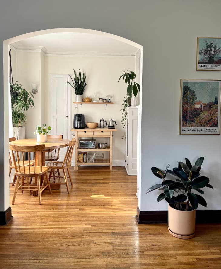 view into dining room with arched doorway wood round table with wood chairs, wood floors, cream walls, kitchen island with coffee maker, toaster oven, floating shelf above with plant, teapot, coffee accessories