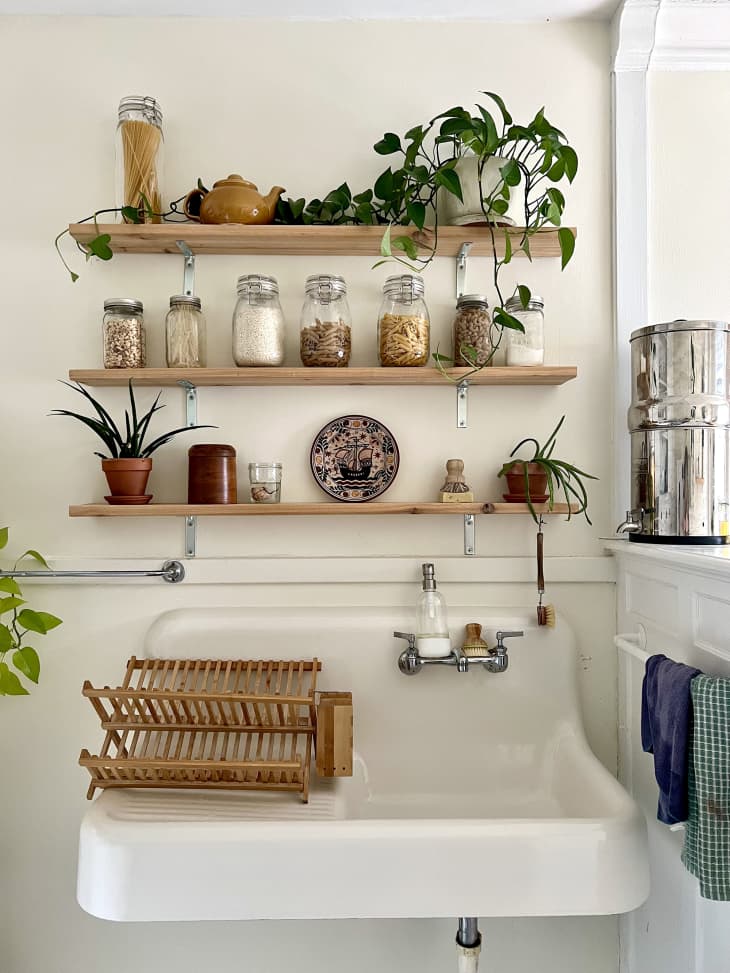 detail of narrow wood shelves on kitchen wall with jars of staple foods, plants. white sink below with wood dish rack