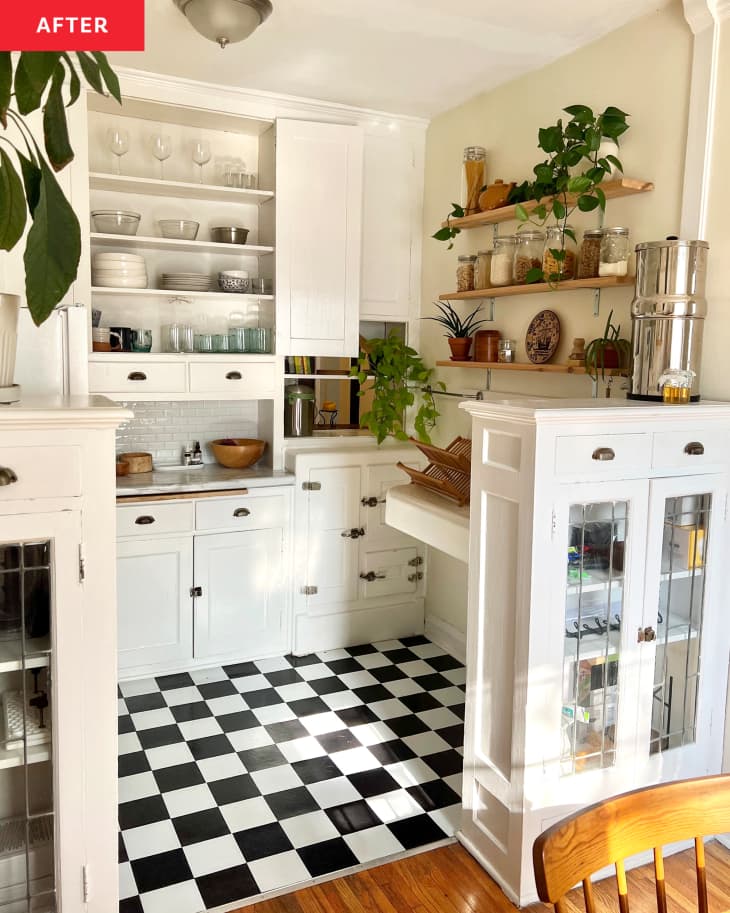 kitchen with white cabinets with black pulls, black and white checkered floor, narrow wood wall shelves with food storage jars and plants, small built in extra counter space, buff colored wall, white cabinet with glass doors, main kitchen cabinet open to show white shelves with glasses and dishes