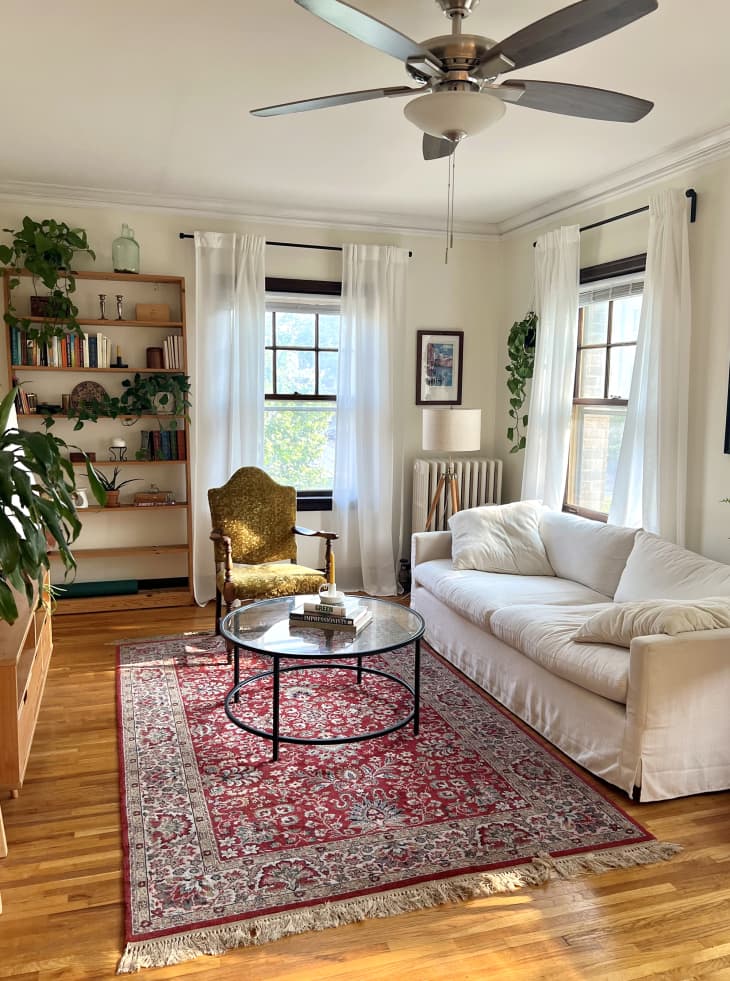 Living room with white sofa, red patterned area rug, round glass top coffee table, 3 windows with sheer white curtains, tall wood bookshelves with books, plants, decor, antique accent chair, ceiling fan
