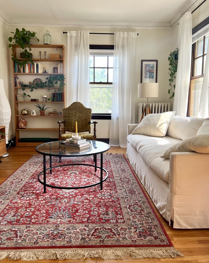 Living room with white sofa, red patterned area rug, round glass top coffee table, 3 windows with sheer white curtains, tall wood bookshelves with books, plants, decor, antique accent chair