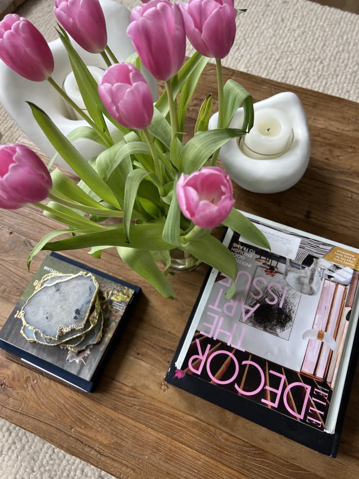 detail of wood coffee table with a decor magazine, white candle holder with candle, vase of pink tulips, stack of coasters