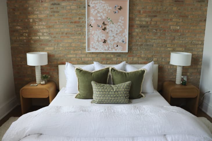bedroom with bed with white bedding and green velvet throw pillows, matching midcentury modern nightstands with white lamps. Exposed brick wall with framed butterfly art