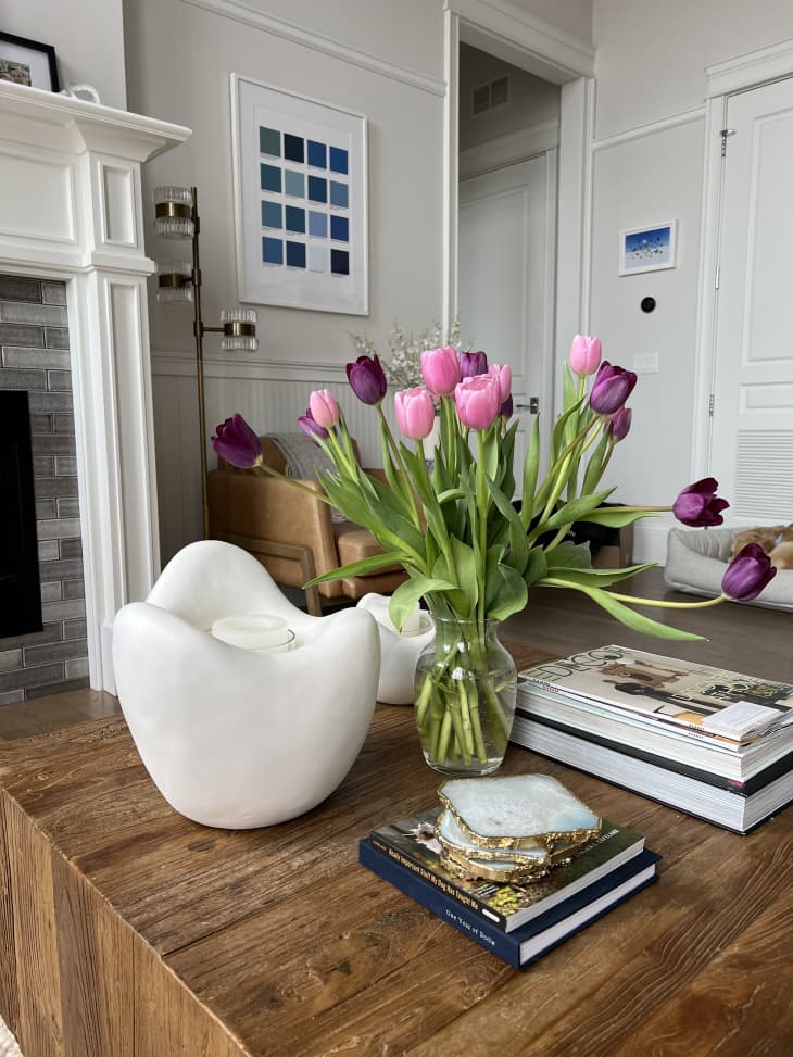 wood coffee table with a decor magazine, white candle holder with candle, vase of pink tulips, stack of coasters. gray brick with white trim fireplace can be seen behind. Walls are pale gray with white trim