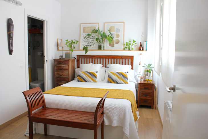 bedroom with white walls, bed with mustard, gray and white bedding with wood headboard, wood side tables, shelf over bed with plants, art, wood slat bench at foot of bed, wood floors