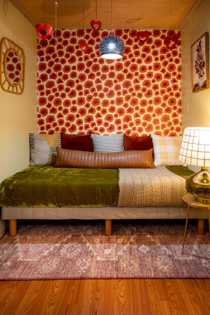 view into basement bedroom with cozy bed with green velvet bedding and red pillows, red and white floral wallpaper wall behind, wood floor, some wall art that matches wallpaper