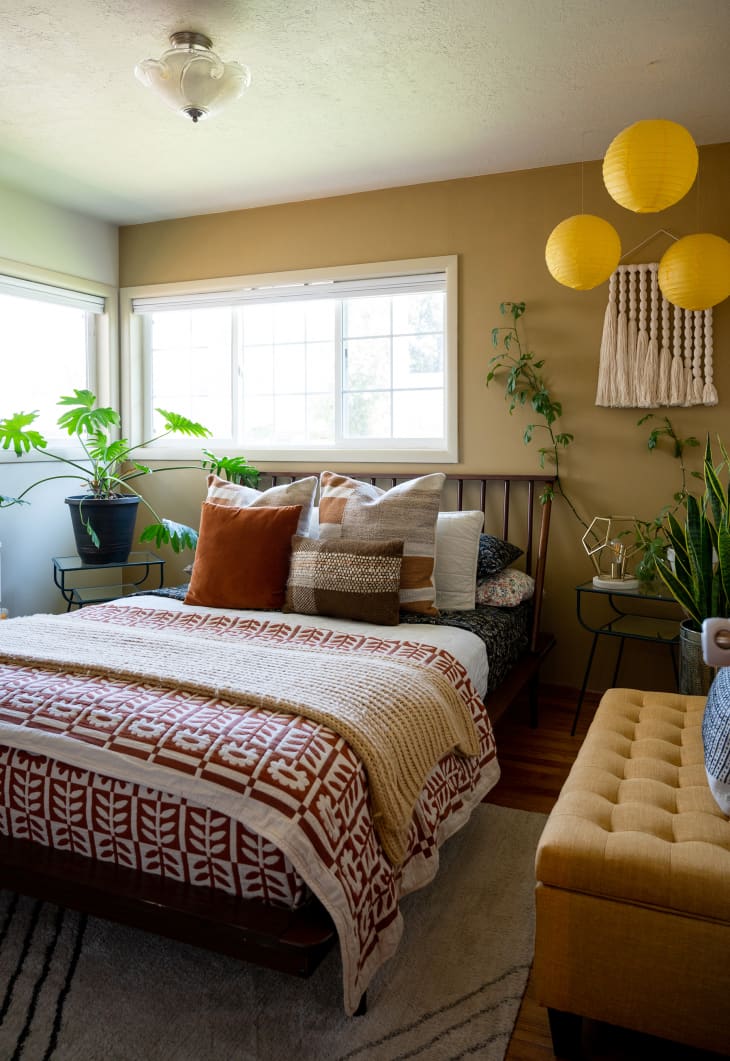 bedroom with one ochre accent wall, warm colored patterned coverlet and navy patterned sheets, and pillows, window behind bed, hanging yellow paper lanterns, large snake plant on floor