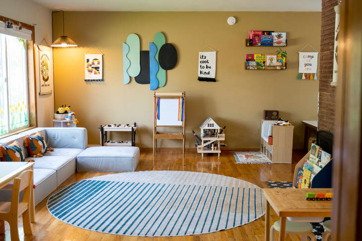 Living/kids classroom area in home. Lots of kids toys, learning materials, fun and inspirational art, muted yellow and green painted walls, a kids' play kitchen, shelves for kids' things, kids' tables with stools, shelves of books, large pale gray sectional with bright throw pillows