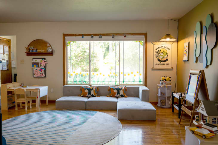 Living/kids classroom area in home. Lots of kids toys, learning materials, fun and inspirational art, muted yellow and green painted walls, a kids' play kitchen, shelves for kids' things, kids' tables with stools, shelves of books, large pale gray sectional with bright throw pillows