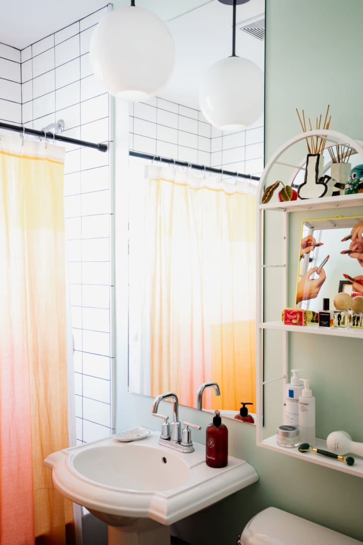 bathroom with shower with white tiles with black grout, pale orange shower curtain, pale sage green walls, shelves with grooming products, reed diffuser, white pedestal sink