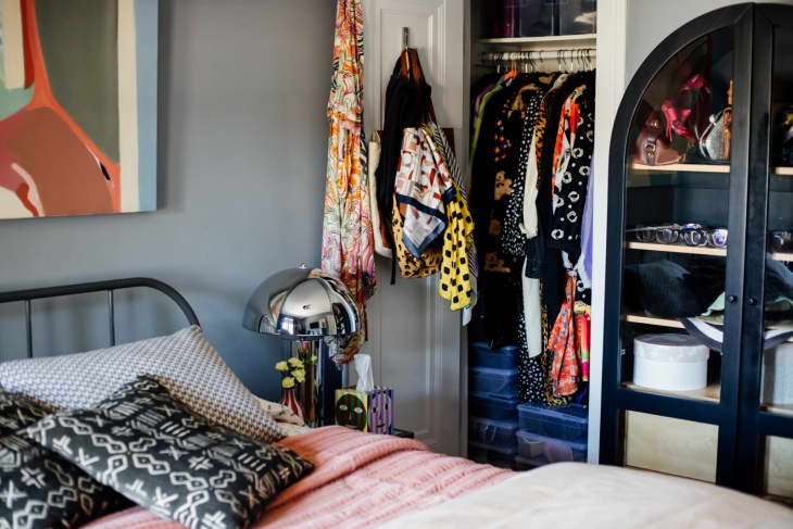 bedroom with gray walls, large abstract painting over bed, black metal headboard, bed with black and white patterned throw pillows, mushroom shaped metal lamp on bedside table, closet open in background with clothes hanging, black and glass arched cabinet with accessories