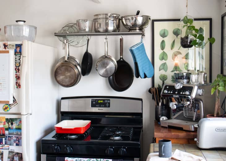 Black stove with pots and pans, oven mitts hanging above on shelf/rack. Espresso machine and toaster on counter to the right
