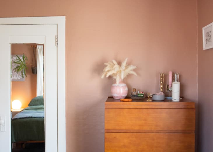 bedroom with peach or pale terracotta walls, bed with pale green bedding, framed art and guitar on wall, arched full length mirror, wood dresser with jewelry, other decor