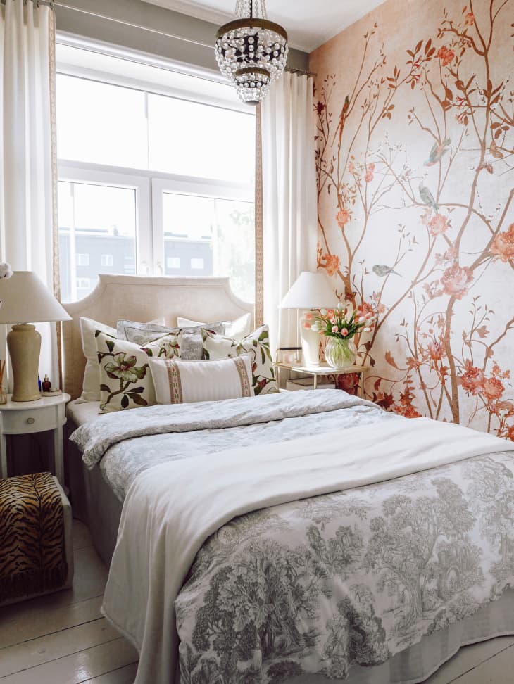 Bedroom with light wood floor, white walls and one accent wall with plants, flowers, birds, crystal pendant light/chandelier, art wall, large window behind bed. Bed has toile style floral bedding