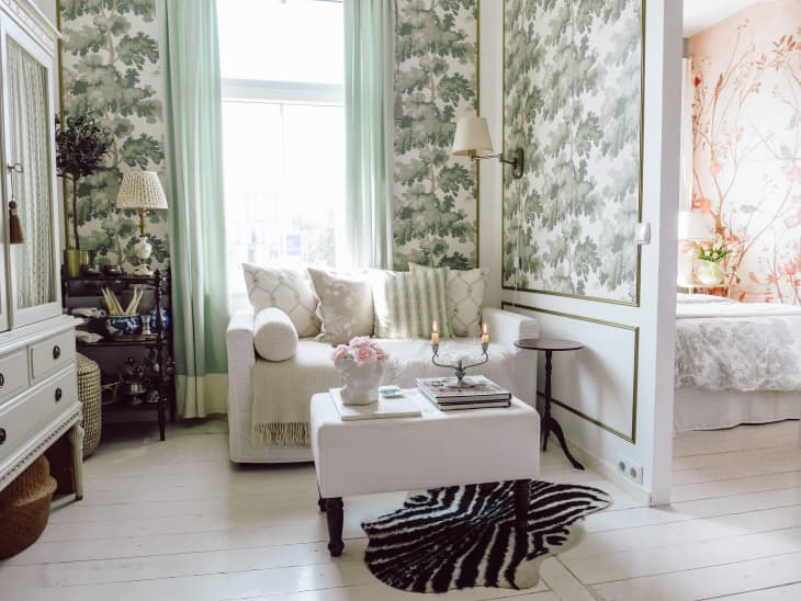 Living room with white sofa, white and green botanical wallpaper, white coffee table with black legs, zebra rug, white wood floor, white armoire, crystal chandelier