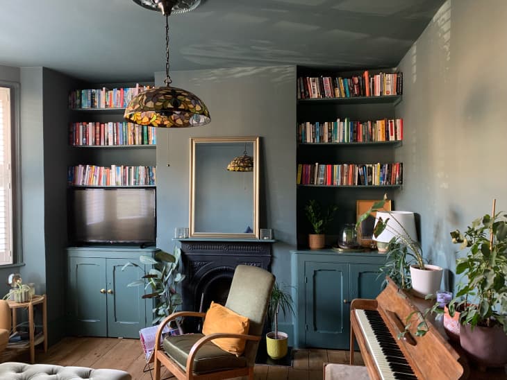 living room or lounge with deep blue walls, bookshelves over TV with blue cabinet under, wood floors, natural area rug, one camel loveseat in window, gray velvet sofa, fireplace, rustic wood piano, tiffany glass style pendant light