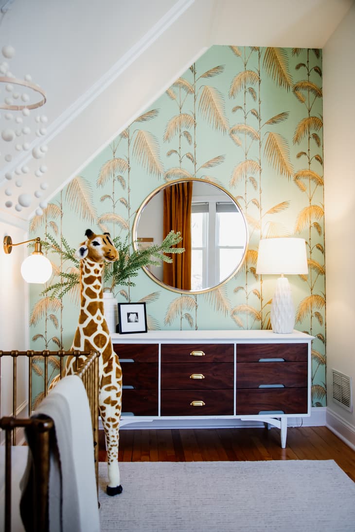 Botanical wall paper in nursery with large stuffed animal giraffe near dresser.