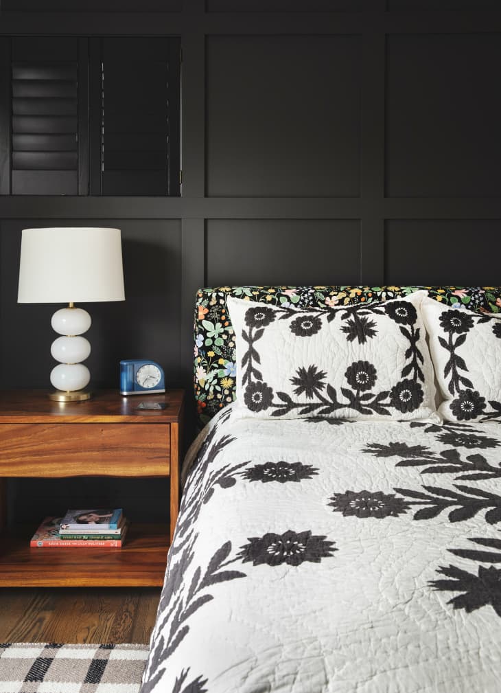 Bedroom with black accent wall behind bed, Bed has black and white floral quilt, pillowcases. modern white lamp on wood bedside table