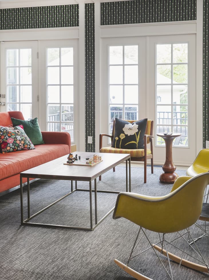 Living room with orange sofa, 2 yellow midcentury rocking chairs and one orange and yellow striped armchair with wood arms and legs, gray/neutral area rug, white walls and large windows with black and green botanical wallpaper accents