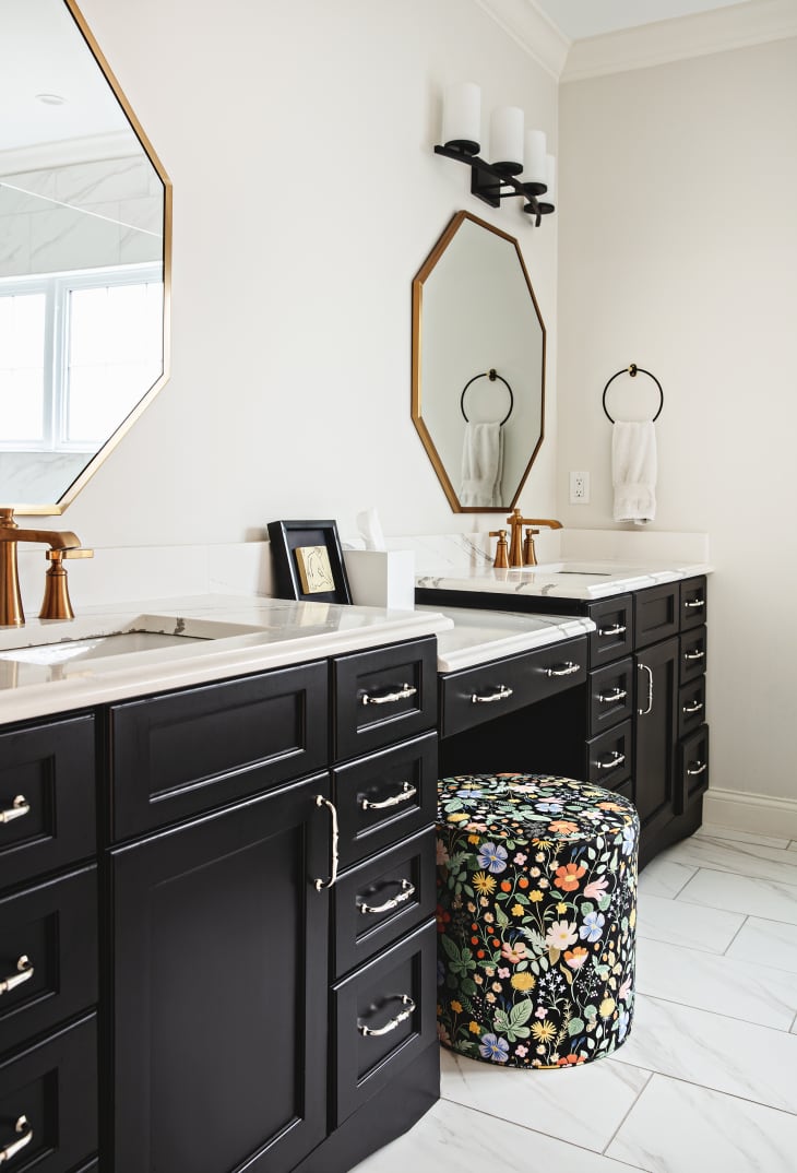 Bathroom sink area with black drawers and cabinets, white marble countertops, brass/copper frame octagonal mirrors, small black floral seat/stool