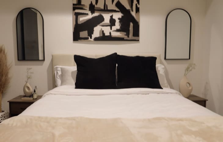 Bed with cream colored upholstered headboard, white bed linens and black pillows. Art work above bed mimics color scheme throughout space.