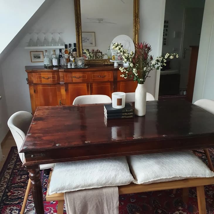 Dining room table during the day with floral arrangement in white ceramic vase and scented candle beside on a stack of books. Bar credenza with a mirror on top sits to the back of the room.