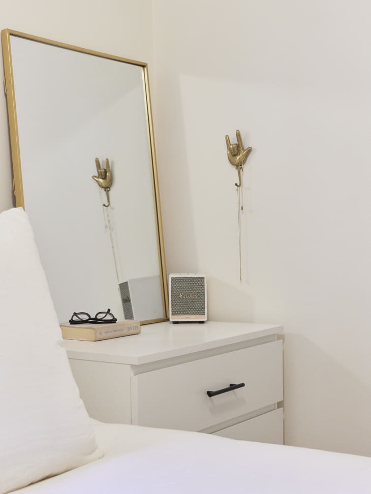 Nightstand corner of an all-white, minimal bedroom with a gold frame mirror on top