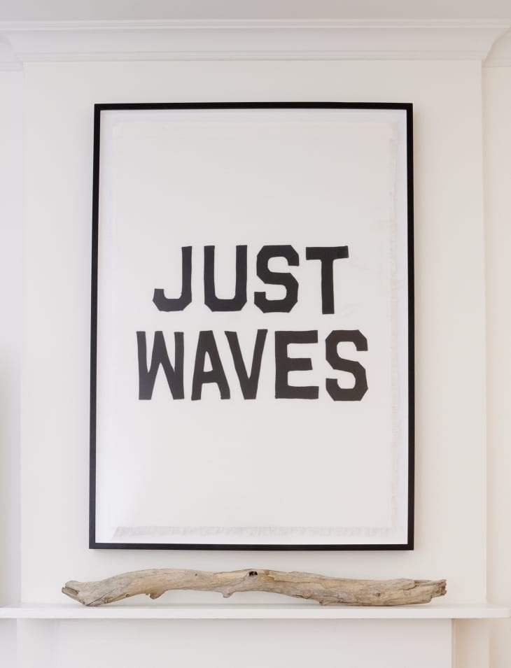 Close-up photo of a minimal white fireplace mantel with one piece of driftwood on top, with a large black and white framed print above that reads "JUST WAVES".