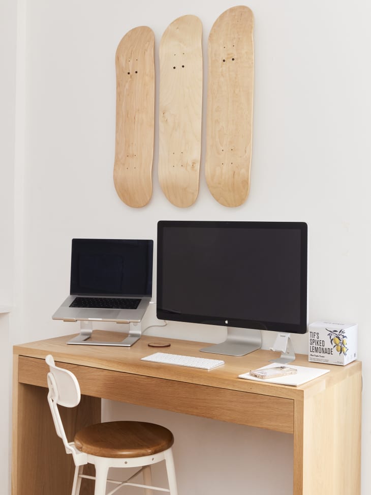 Minimal and modern natural wood desk with Apple laptop and monitor on top, with three natural wood skateboards hanging on the wall above.