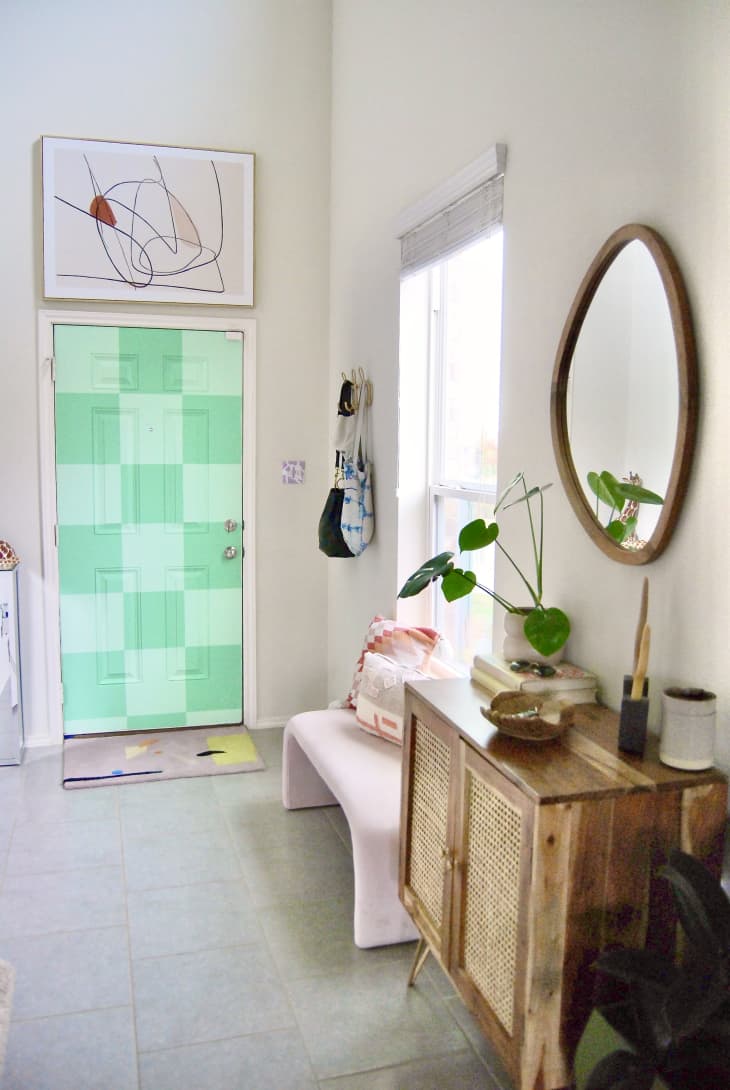 Colorful entryway with monochromatic checkerboard painted door. Abstract artwork above entry door and bench in hallway. Wooden sideboard with caning detail and asymmetrical mirror above sideboard.