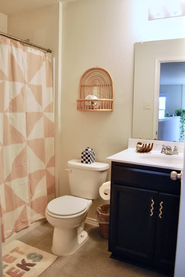 Small bathroom with black vanity with wavy handles and white surface top. Pink and white shower curtain in bathroom and wooden shelf above toilet.