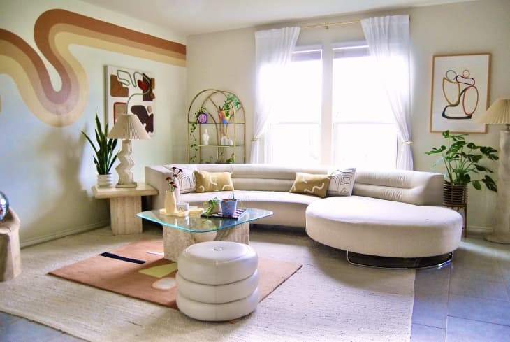 Monochromatic living room with wavy coffee, tan and cream colored decal on wall. Hand sculpture holding disco ball near wall. Cream colored sofa with glass coffee table in the center and light colored leather ottoman in room.