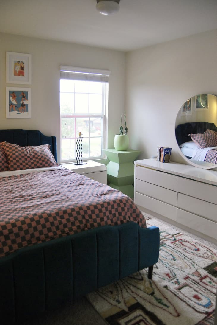 Colorful bedroom with velvet upholstered bed 
made with checkerboard bedding. White glossy dresser topped with round mirror and green decorative stand are across from bed.