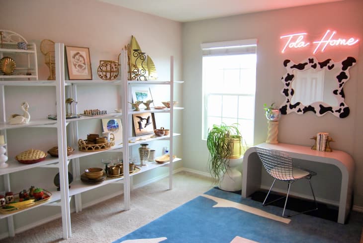 Home office with neon signage above desk and blue abstract rug on the floor. White shelves filled with decorative objects line the wall.