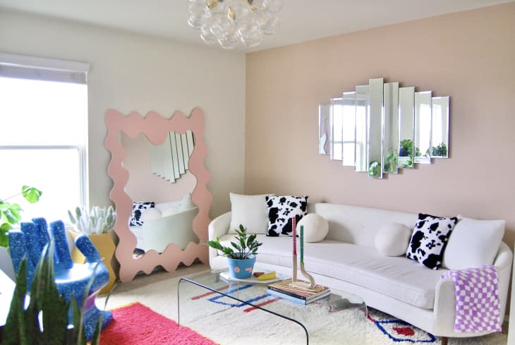 Colorful sitting room with bubble chandelier and wavy pink standing mirror in the corner. Blue hand chair sits across from glass coffee table topped with colorful taper candles and potted house plant.