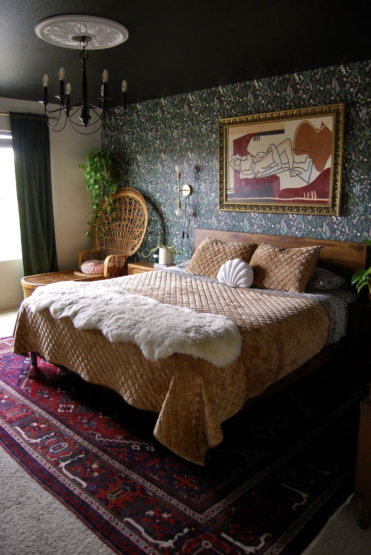 Bedroom with green floral wallpaper and dark painted ceiling. Wicker chair in corner beside bed. Bed is made with tan velvet blanket and animal print linens. Red and blue rug lines floor.