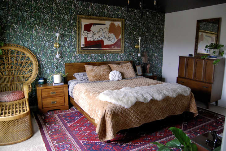 Bedroom with green floral wallpaper and wicker chair in corner beside bed. Bed is made with tan velvet blanket and animal print linens. Vintage wooden dresser across from bed.