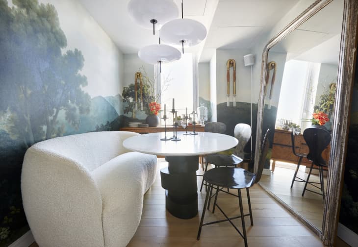 Round white and black dining table in dining room with landscape mural wall. Black chairs, and a cozy white banquette that's more like a sofa