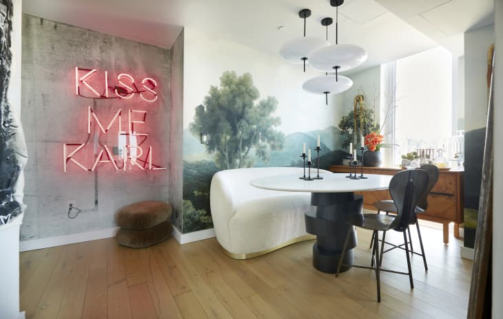 View of dining room and into hall. Round white and black dining table with landscape mural wall. Black chairs, and a cozy white banquette that's more like a sofa. Neon sign in hallway reads "Kiss me Karl"