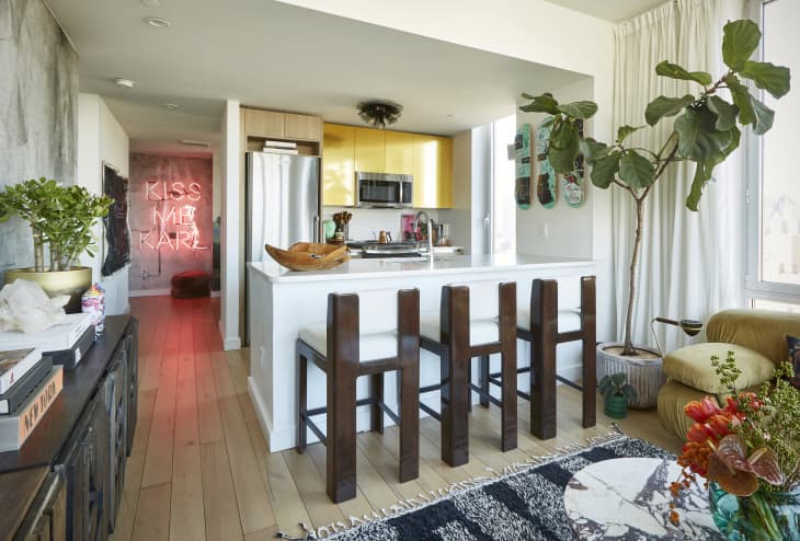 View into kitchen with barstools, large fiddle leaf fig plant. View down the hall, neon sign saying "Kiss Me Karl"