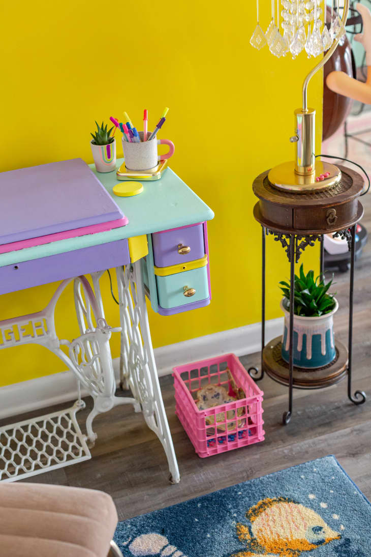 detail of desk area. Bright yellow wall, colorful desk, small round tiered table with lamp and plant