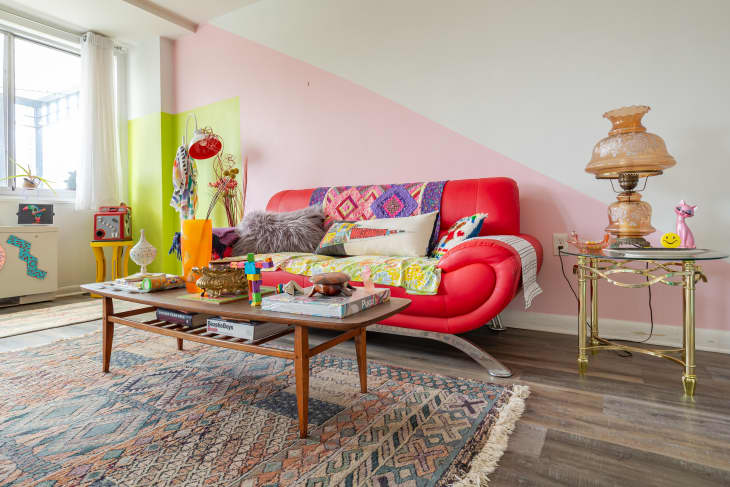Colorful living room with red sofa, patterned throw pillows, large windows, pink, white, chartreuse, and lavender walls,  wood accent chair with shag throw, wood coffee table with books and colorful design objets