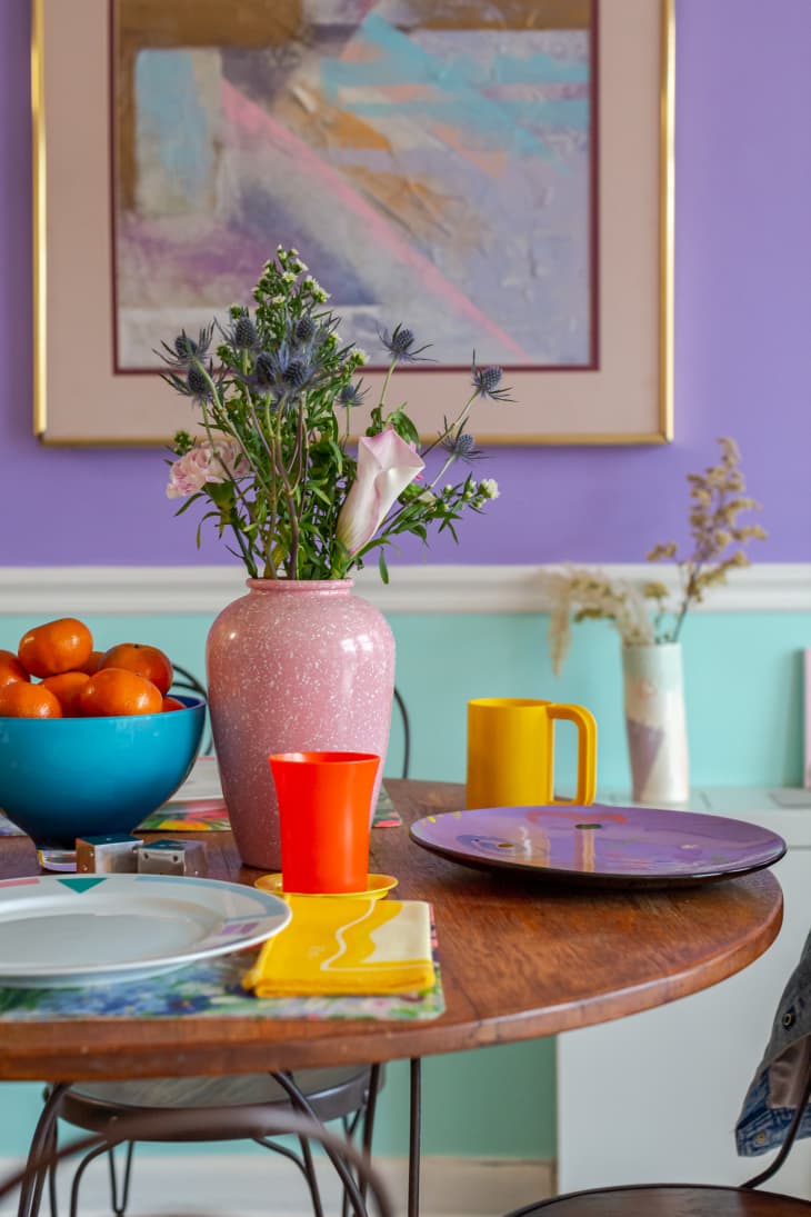 Dining table with lyrical iron chairs, walls painted in color blocks of pale aqua, lavender, multiple vases, colorful plates, bowl of tangerines