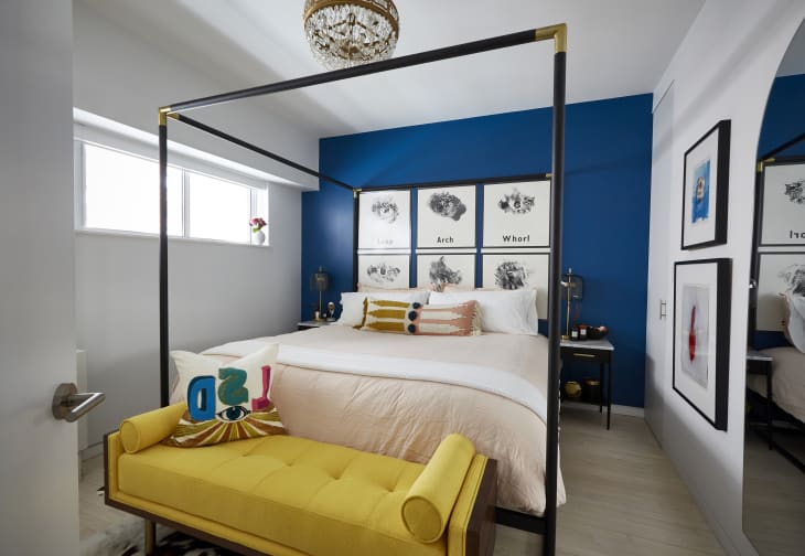 bedroom with one blue accent wall behind canopy bed, gallery wall, yellow settee at foot of bed