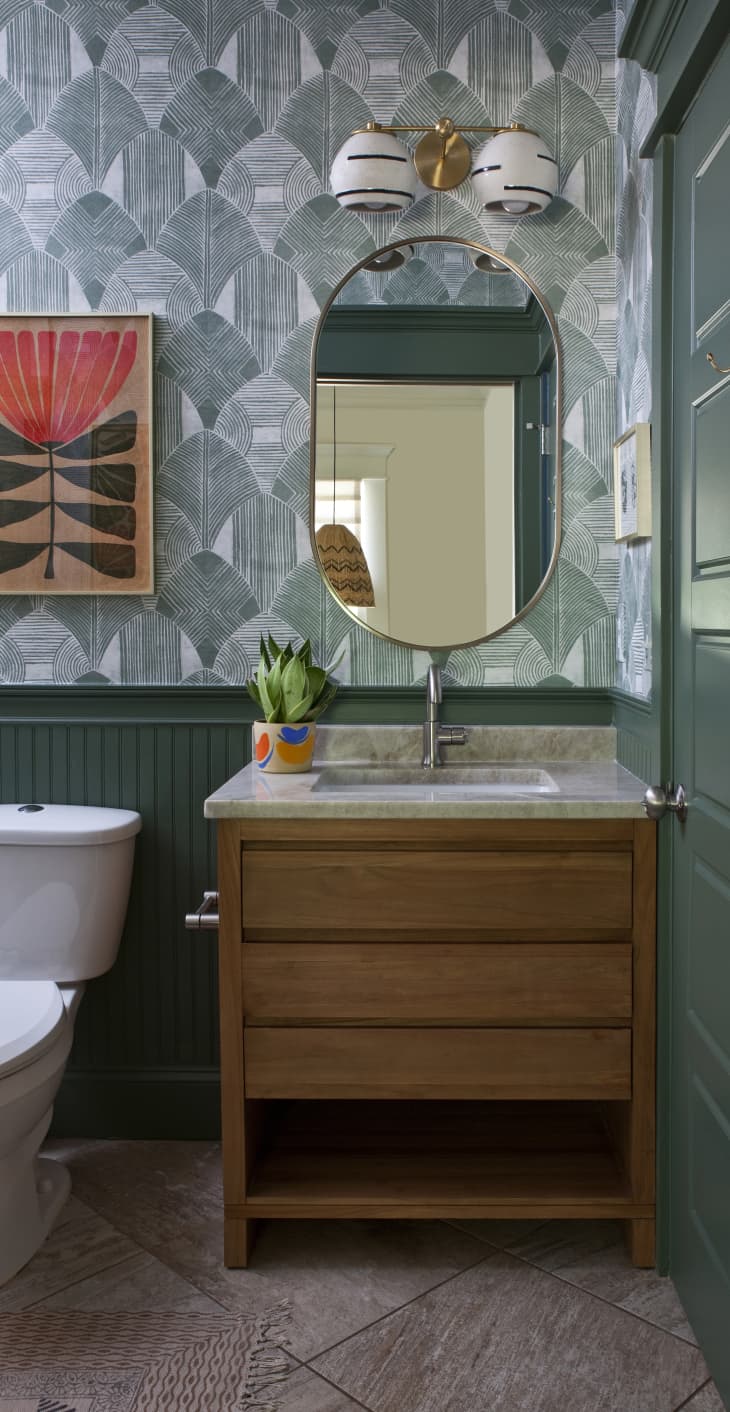 Bathroom after makeover. Patterned wallpaper, black and white and gold wall sconce, oval mirror, wood sink cabinet, green painted door and wainscotting