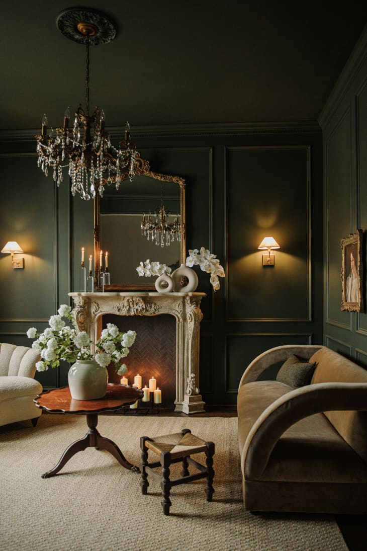 Living room with dark green walls, chandelier, ornate fireplace with candles, natural textile rug, European style furnishings