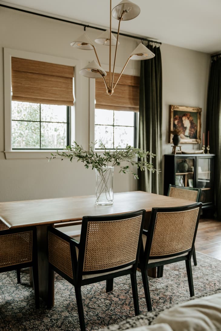 View into dining area. Wood dining table with rattan and black chairs, windows with brown shades and floor to ceiling green curtains, elegant modern chandelier.