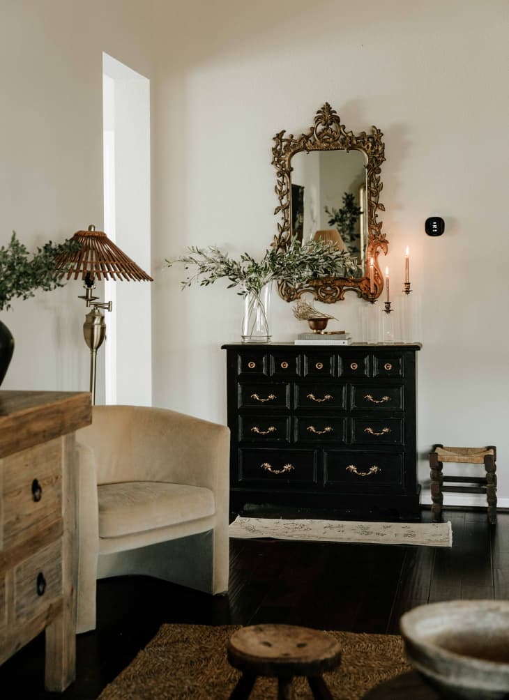Antique black chest of drawers with large ornate bronze mirror above. Vase with olive branches and candles on dresser. Small stool on floor next to it